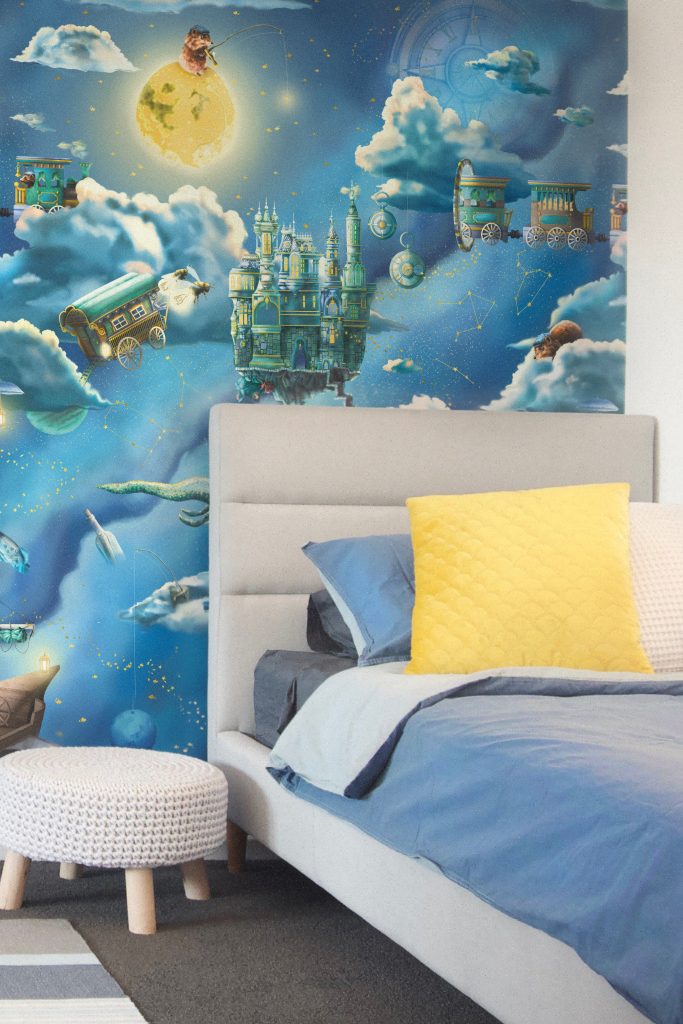 Kids galaxy interior wallpaper design. Features a night sky space theme with moon, clouds, dragon, compass, fishing and fairy tale creatures. In blue colours styled with a grey bed and yellow cushion.